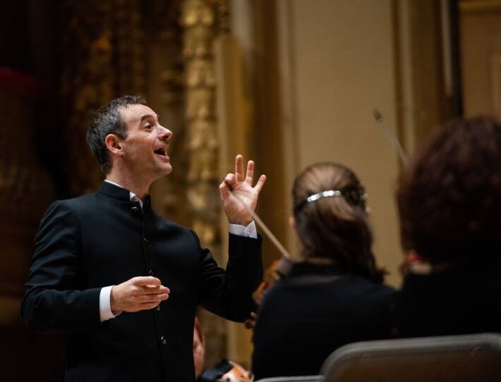 Jonathan Cohen conducts the Handel and Haydn Orchestra. He is wearing a black dress jacket and white shirt. He is smiling. His left hand is raised as he address the musicians.