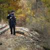A police officer carrying a gun stands alongside a railroad track amid fall foliage