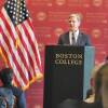 A man in a suit stands behind a podium labeled "Boston College." People are seated facing him.
