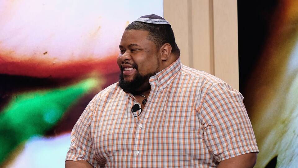 A man wearing a yarmulke and checkered shirt smiles as he sits on stage.