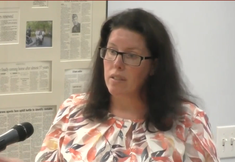 A woman speaks into a microphone with a newspaper clippings displayed on a wall behind her.