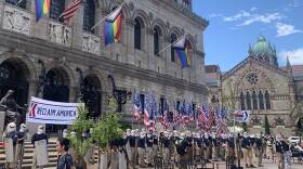 A group of men in khaki pants and face masks hold up American flags and a sign that says "Reclaim America." They stand in front of the Boston Public Library.