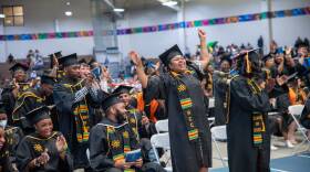 This is a photograph of a college graduation ceremony.  Inside a gymnasium, dozens of college graduate.  donning black robes and mortar boards, applaud and stand amidst of a jubilant moment.