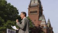 A man in a suit holds a piece of paper with notes as he speaks into a microphone on Harvard's campus.
