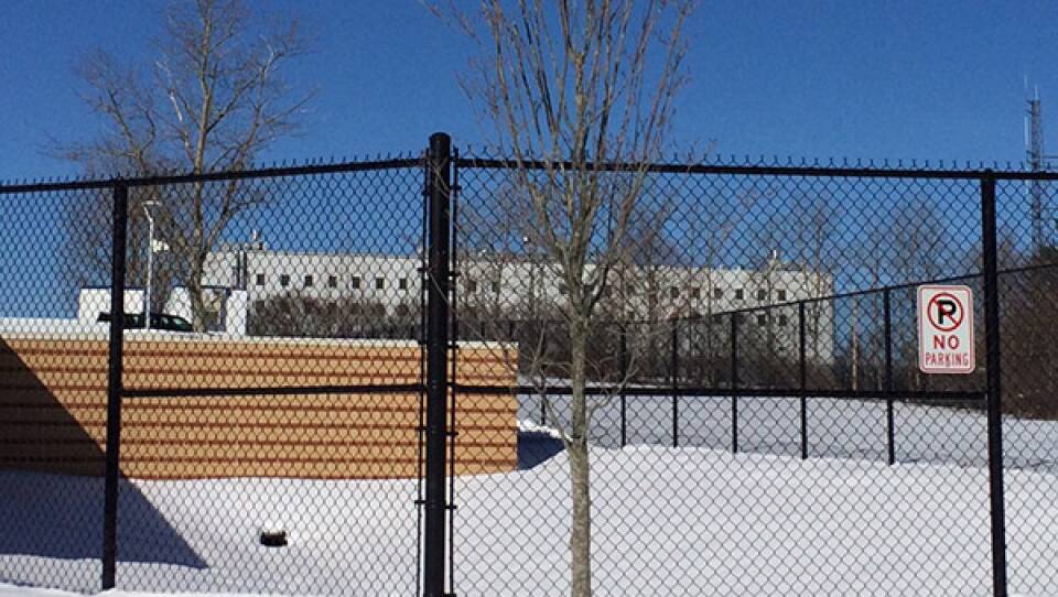 A black chain-link fence stretches across a snowy field in front of a large gray jail building.