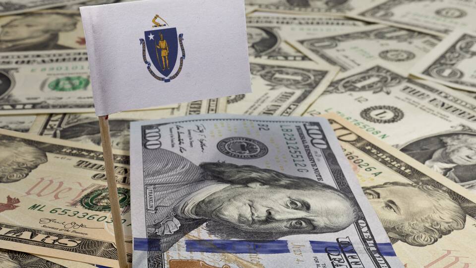 A miniature Massachusetts flag stands erect in a field of U.S. dollars.