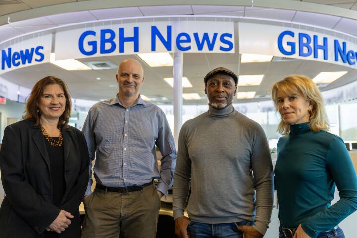 Four people — two men flanked by two women — stand in an office underneath signs reading "GBH News"