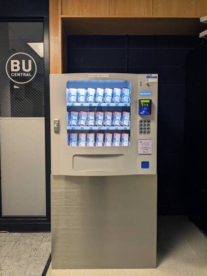 A vending machine with packets of medication sits in a hallway. 'BU' is on the door next to it.