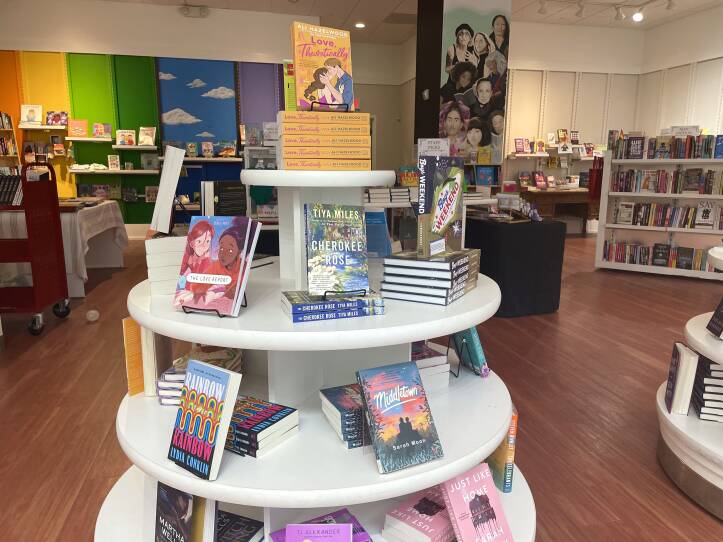 This is a photograph of a bookstore. In the foreground is a round, multitiered book display, featuring new releases. The books are in horizontal stacks with some books propped up vertically. In the background are bookcases. The space is bright. Against the back wall are vertical panels of bright colors, which look like the rainbow pride flag
