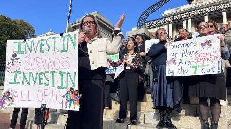 A woman with blond hair and glasses speaks into a microphones. Standing around her are people with signs showing support for funding programs for domestic violence survivors.