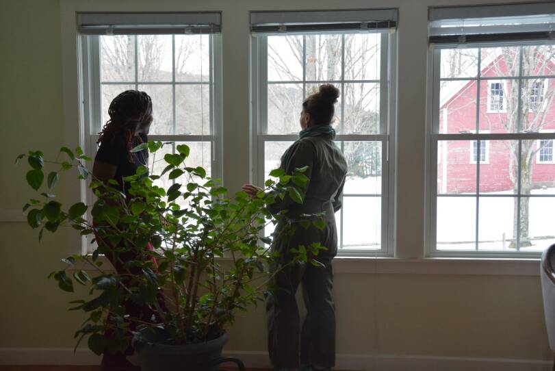 Two women stand in front of a large window that looks out at snow and a red barn. A large green plant is in the foreground.