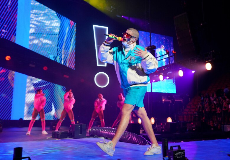 Bad Bunny wears a blue and white two-piece outfit as he walks across a stage. Four back-up dancers are lit in pink behind him as his sings into a microphone he holds up to his mouth.