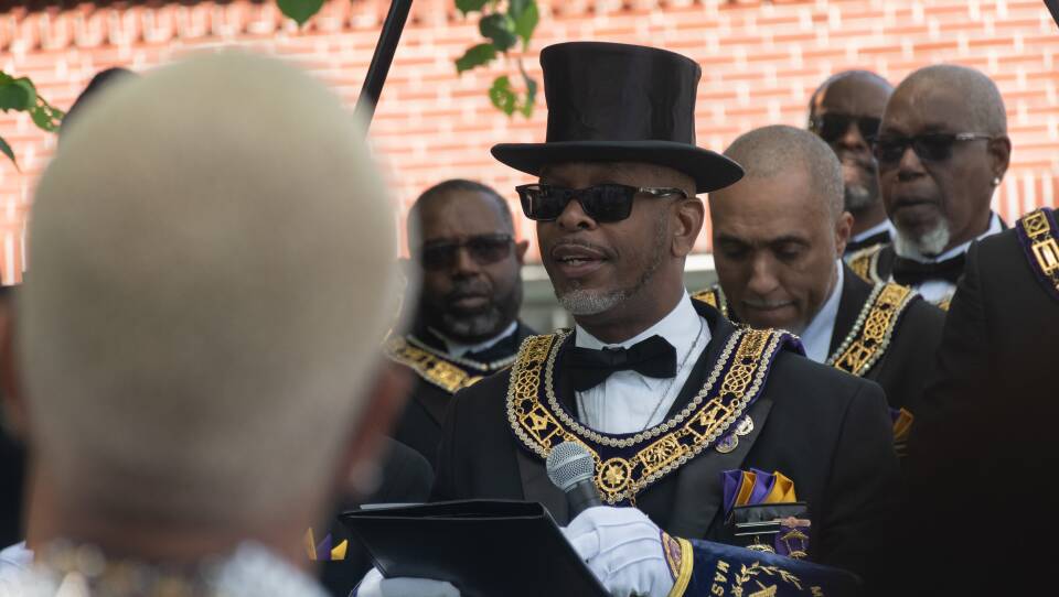 A Black man wearing sunglasses and dressed in Masonic attire -- a black top hat, black tuxedo, jeweled collar -- speaking into a microphone.