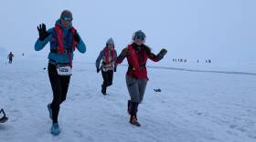 Three individuals in a snowy environment are wearing cold weather clothing and running past the camera.
