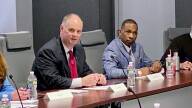 Two men sit at a conference table with a microphone between them during a roundtable discussion. There are water bottles and name placards scattered across the table tops.