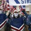 Three people wearing white face coverings, baseball caps and holding metal shields stand in front of a crowd on a city sidewalk.