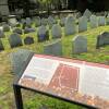 A welcome sign describing the King's Chapel Burying Ground sits among the rows of ancient tombstones. The sign is partially obscured by rows of several hundred pennies left by visitors.