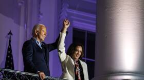 President Biden and Vice President Kamala Harris join raised hands and smile looking out from the White House balcony.