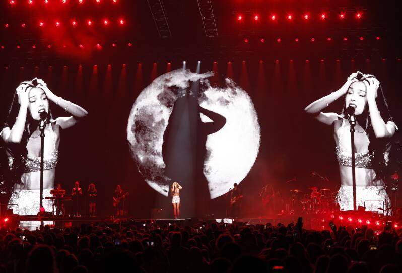 Projected screens behind Olivia Rodrigo show her intensely singing into the microphone on stage. An image of the moon is cast on her.