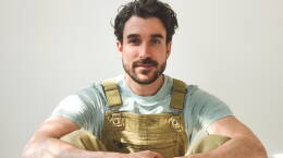 AJR's Adam Met sits on the floor. He is wearing brown overalls and a teal shirt.