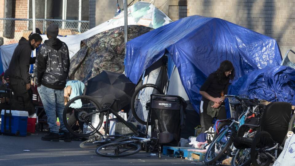 Several tents covered in tarps line the sidewalk. Two men in sweatshirts are standing and talking, and another woman stands nearby smoking a cigarette.