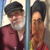A man with a white beard and black hat stands next to a wooden statue of the artist Frida Kahlo.