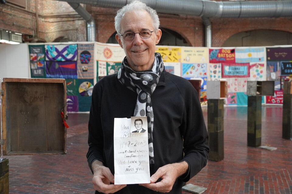 A man stands in the middle of an art installation. He is holding a memorial to his friend who died from complications from HIV/AIDS, which is part of this art exhibit. The memorial is a white paper bag and it has a black and white photo of his friend attached to it. On the bag is a handwritten message that says " John Pfisterer 1947-2003.....we love you and miss you"