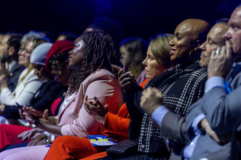 A row of people in an audience have their hands raised and clapping in applause.