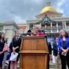 Warren and other leaders stand somberly stand in front of the State House. Warren speaks from a podium.