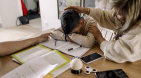 A mother consoles her son as he buries his head in his arms on a table.
