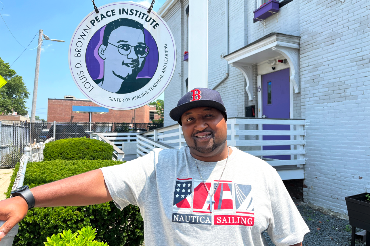 A man smiles for the camera, wearing a baseball cap and T-shirt in front of the nonprofit where he works
