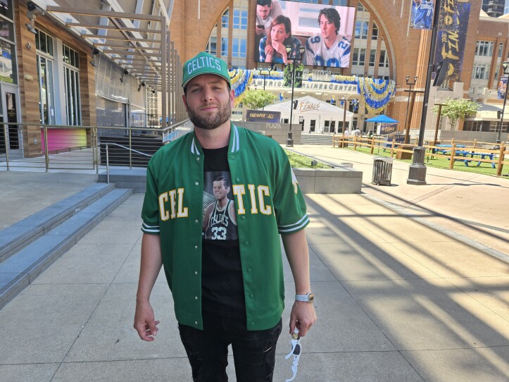A man stands in a green Celtics jacket in front of an arena. He has a green Celtics hat and a black shirt on.