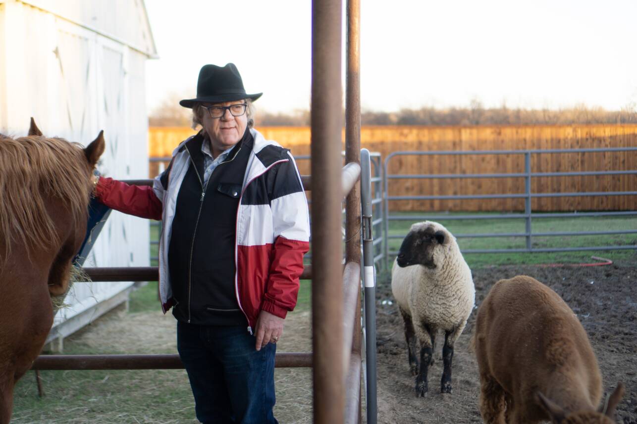 An older man with glasses and a ten-gallon hat pets a horse's head while looking out over a pen with two sheep.