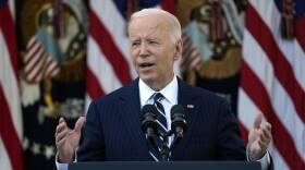 Joe Biden gestures with both hands as he delivers a speech in front of a display of American flags.