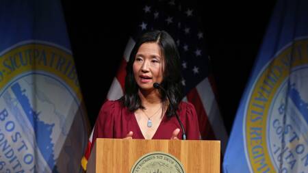Michelle Wu speaks from behind a lectern on stage, with Boston city flags hanging behind her.