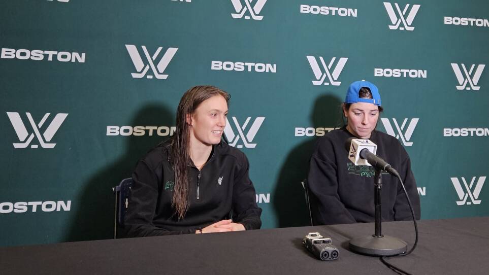 Two women sit in front of a microphone. Behind them is a green and white background with a logo featuring two hockey sticks and the word Boston.