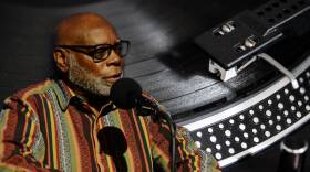 A Black man in a brightly striped shirt is speaking into a microphone. The image is overlaid on an image of a record player.