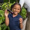 A young child in a blue shirt and braids holds up an eggplant and smiles.