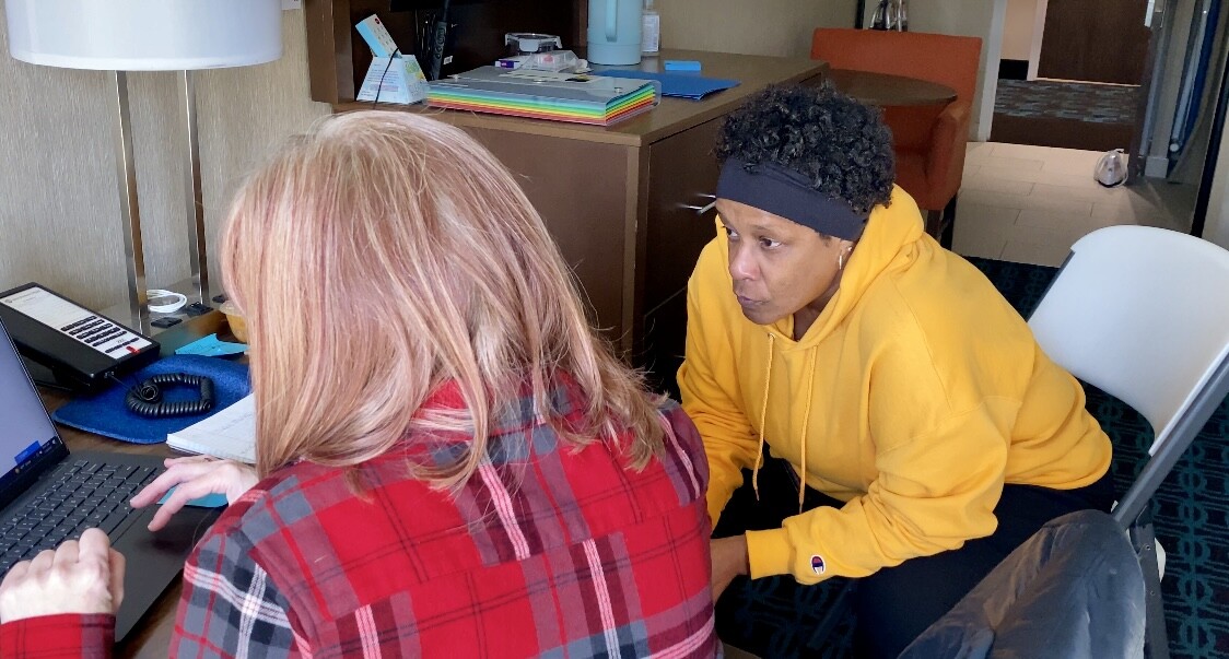 A middle aged black woman sits with a woman with red hair and both look intently at a computer screen as they search for housing for Susan.
