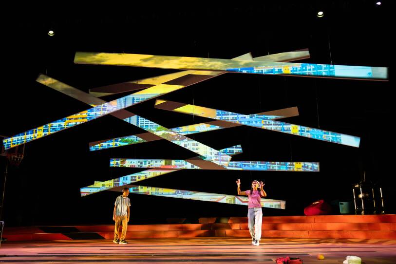Two children stand on a large stage below large, suspended props of colorful, horizontal blocks stretching up to the top of the stage.