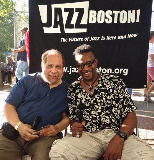 This is a photograph of Eric Jackson and Dr. Bill Banfield. They are at a Jazz Boston event, sitting outdoors. It is a sunny day and it appears to be warm as they are both wearing short sleeved shirts. They are both holding microphone. The background is a large vertical banner that says "Jazz Boston! The future of jazz is here and now."