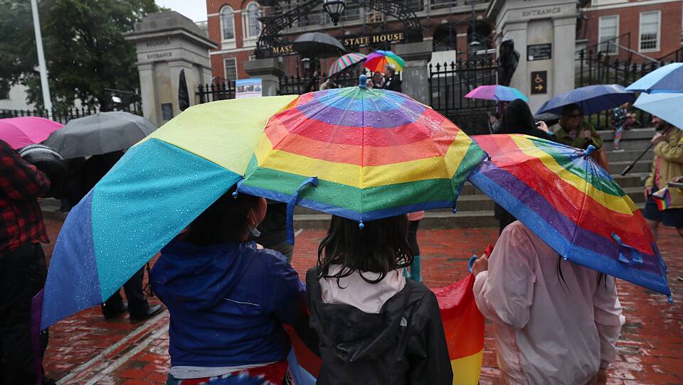 People holding rainbow umbrellas stand on the cobblestone walkway in front of the Massachusetts State House.