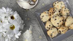 Table with a cup of coffee, a vase of white flowers, and a square plate filled with mounded, cakey chocolate chip cookies