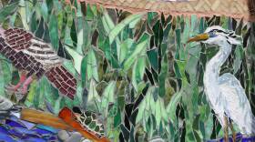 A mosaic of green, blue, white, red and brown glass and stones form an idyllic river scene with a white heron standing in the water.