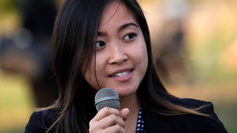 An Asian American woman holds a microphone. She wear a black jacket and looks off to the side.