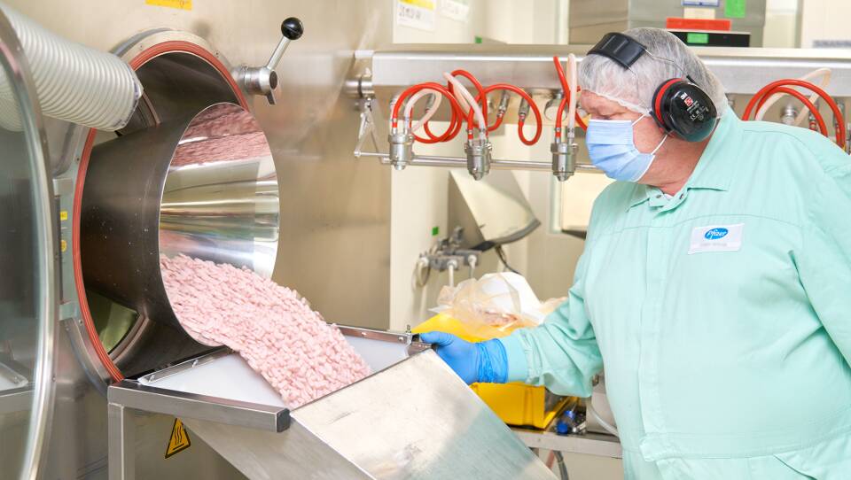 A worker in protective clothing and headphones holds the side of a metal chute as pink pills cascade into it.