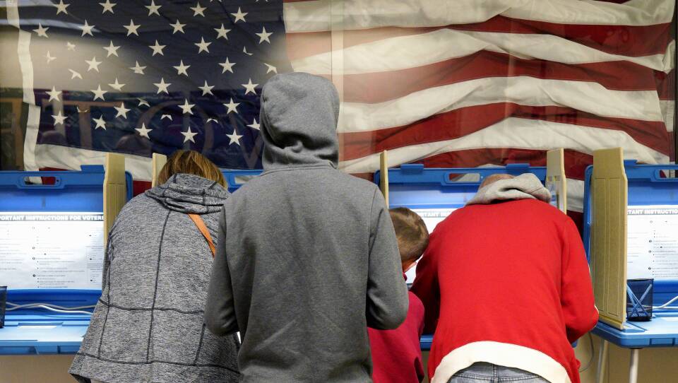 Two adults lean over at voting booths to cast their ballots. A teenager and younger child are standing in between them.