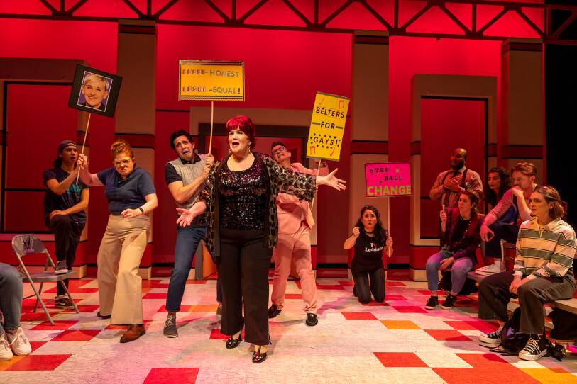 Onstage at the Speakeasy Stage Company, the company perform a musical number during what appears to be a protest in favor of gay rights. signs read "lesbi-honest lesbi-equal!", "belters for gays!", and one features just a photo of Ellen