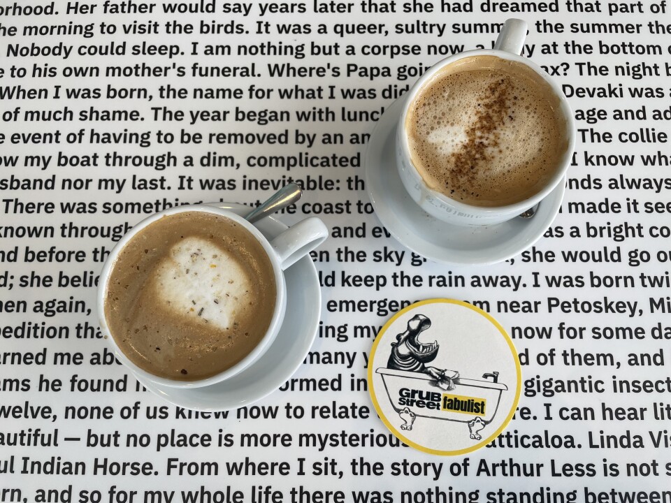 Two cups of cappuccino are on a table that is white, with black writing across it, as if it were a page from a book