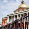 The sun shines bright on the golden dome of the Massachusetts State House, which stands behind an iron fence.